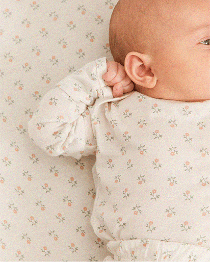 Baby wearing white long-sleeve onesie with small pink and green floral pattern lying on matching floral sheet.