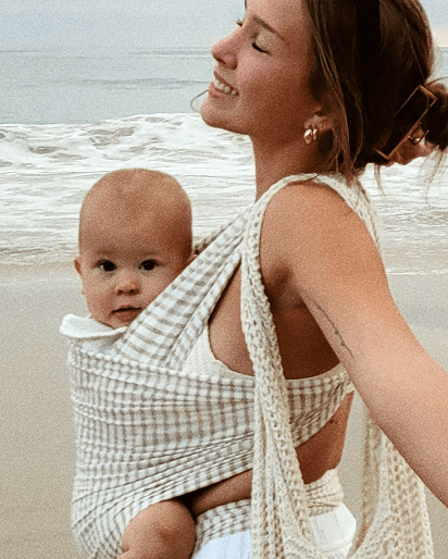 Woman wearing beige and white checkered baby carrier with baby on the beach near ocean waves.