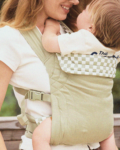 Light green baby carrier with checkered pattern on the upper back panel, worn by a person holding a baby.