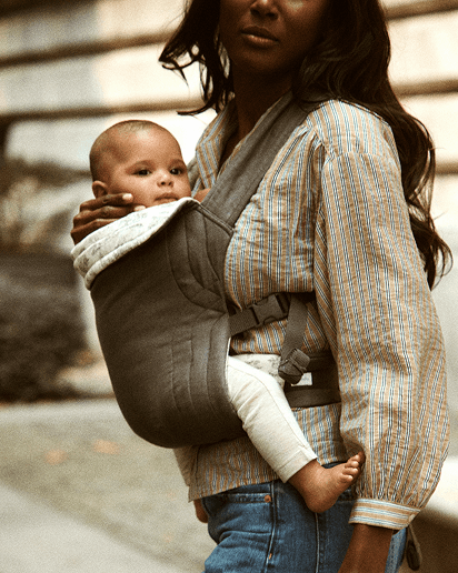 Gray ergonomic soft baby carrier worn by woman holding baby against chest outdoors.