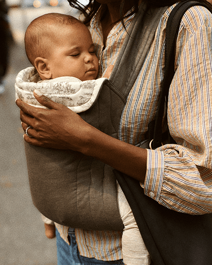 Dark gray baby carrier with white floral padded head support, holding a sleeping baby, worn by a person in a striped shirt.