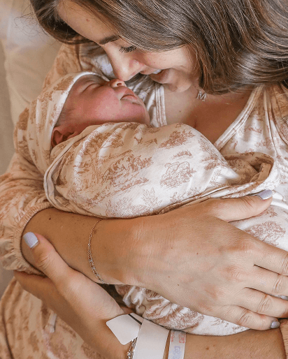 A close-up lifestyle shot of a woman cradling a sleeping newborn baby. The baby is swaddled in a soft, champagne-colored satin blanket with a delicate brown floral toile pattern, and wears a matching patterned swaddle wrap and headband.