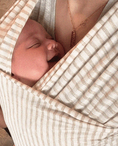 A close-up lifestyle shot of a sleeping newborn baby nestled inside a beige and white striped baby wrap or carrier worn by a mother.