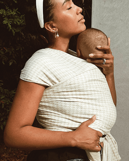 Woman wearing a light beige textured baby wrap sling holding a baby close to her chest, with a white headband and earrings.