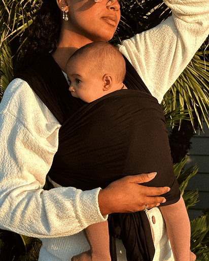 Black fabric baby carrier wrap worn by a person carrying a baby against their chest outdoors with greenery background.