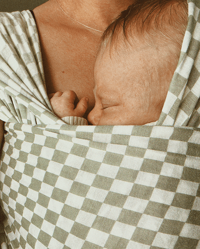 A close-up lifestyle shot of a baby wearing a soft, checkered baby wrap or sling with a mother's chest and shoulder visible.