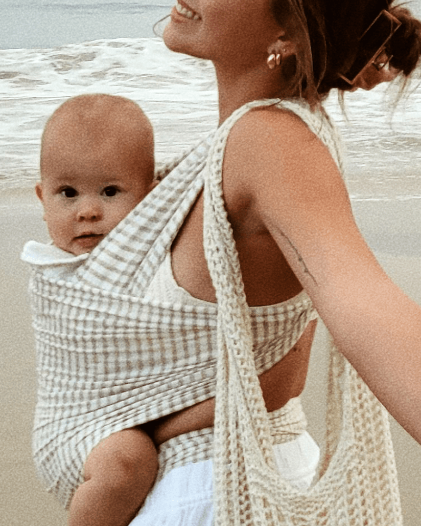 Beige and white checkered baby wrap carrier worn by woman at beach holding baby facing forward
