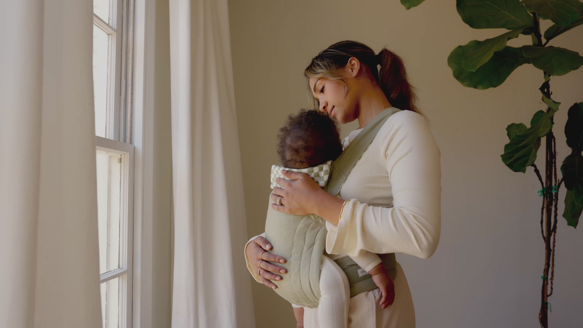 Mom wearing a sage chequer soft structured baby carrier rocking baby back and forth while looking out window with cream curtains flowing in wind
