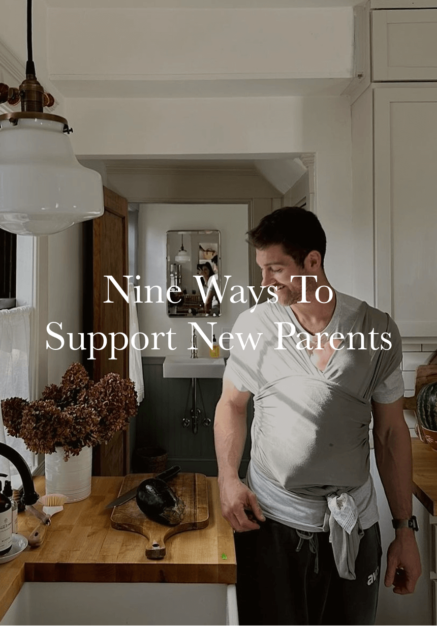 Man in gray shirt and black pants using gray baby wrap carrier in kitchen with wooden counters and dried flowers.