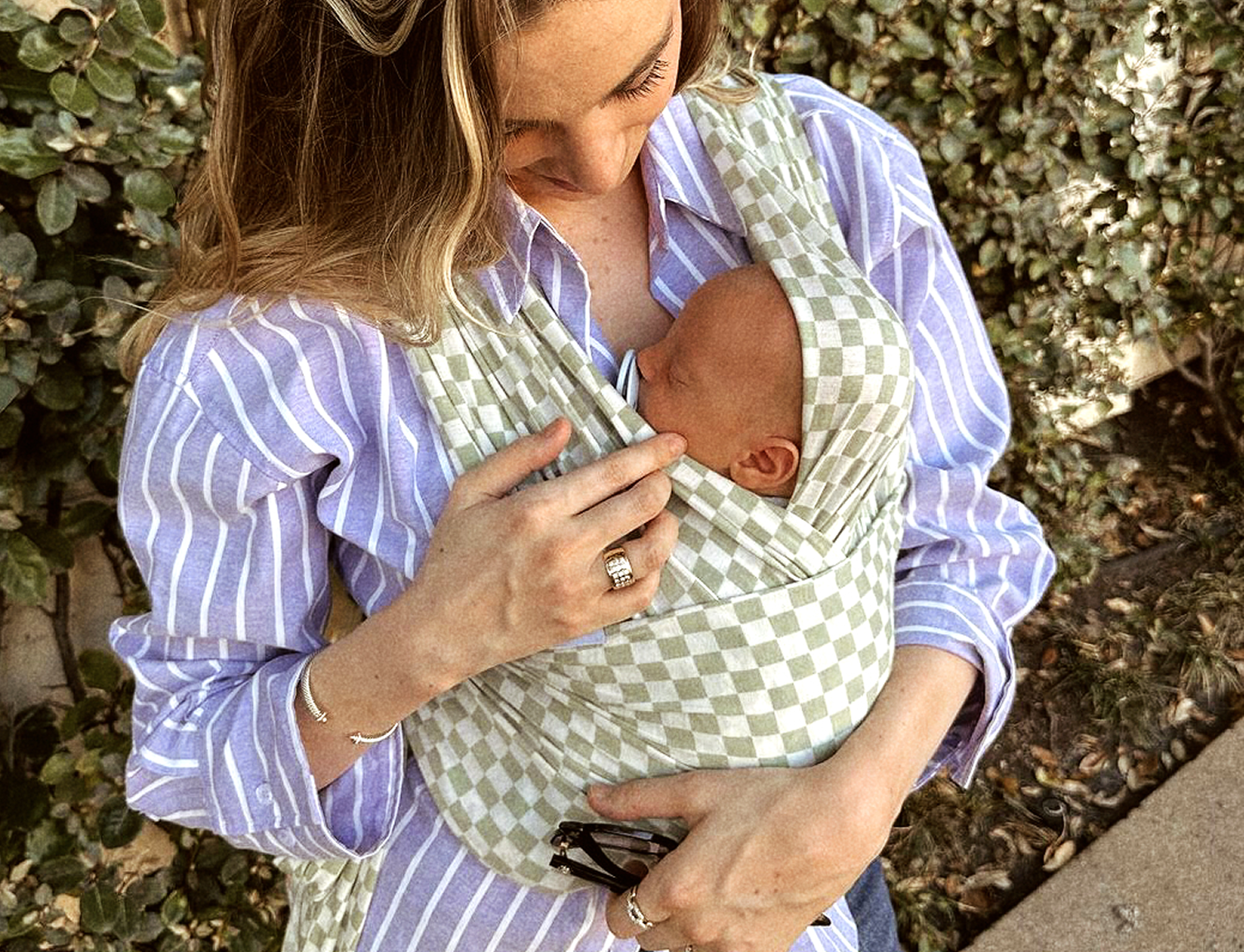 A woman outdoors wearing a light green and white checkered baby wrap carrier shirt, holding a baby nestled inside the fabric.