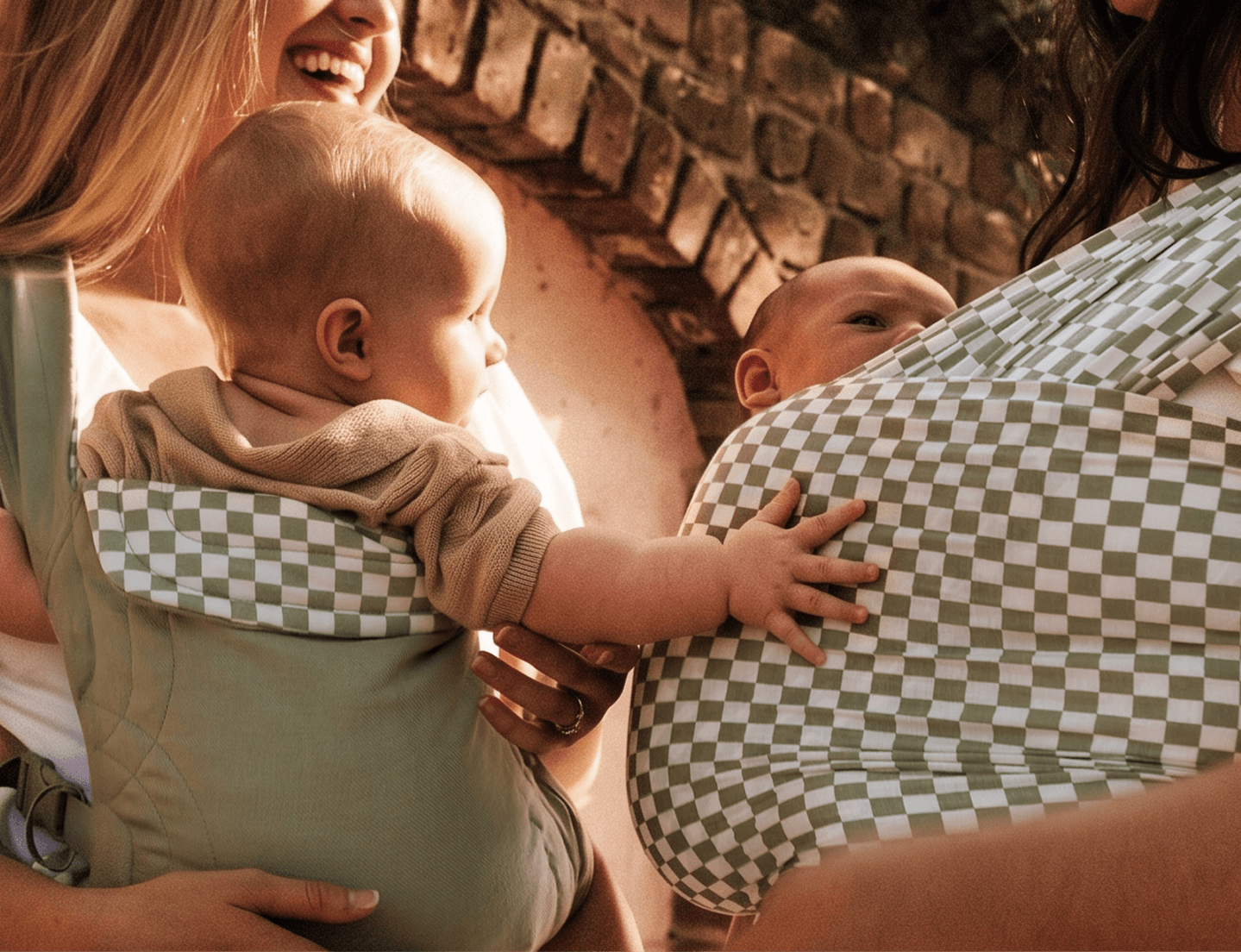 Two babies in carriers, one solid sage green and one green and white checkered pattern, held by smiling adults.