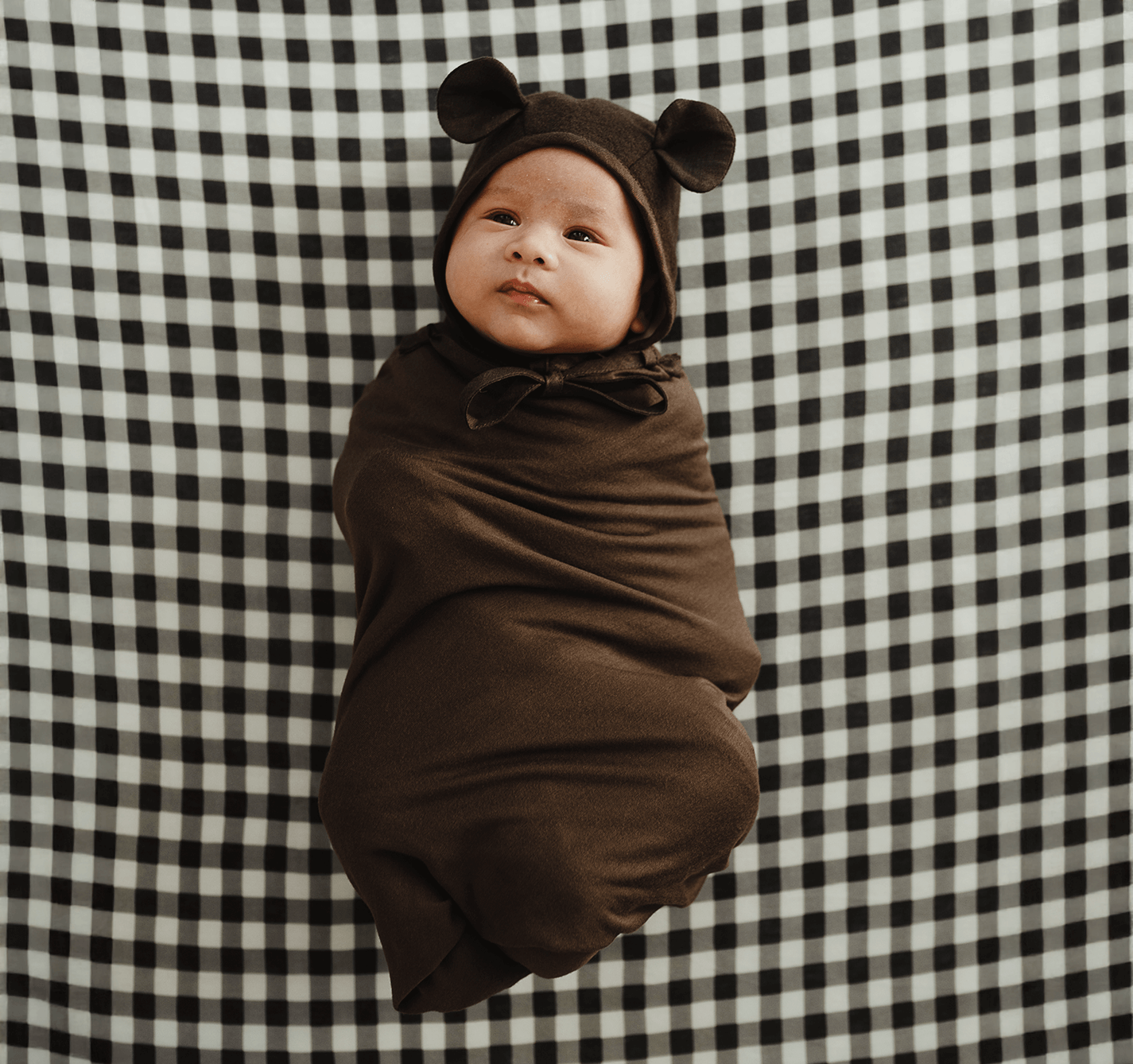 Infant wrapped in dark brown swaddle blanket with bear ear hood lying on black and white checkered fabric.