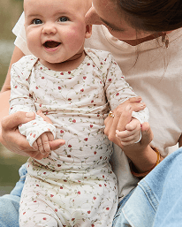 A baby wearing a long-sleeve cream bodysuit with a small red and green floral pattern, held by an adult outdoors.