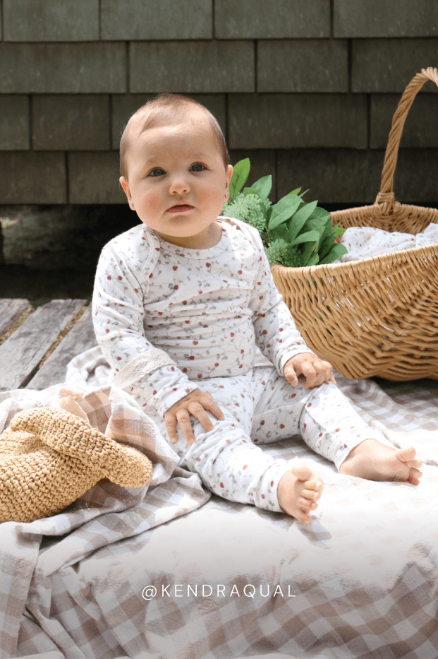 Baby sitting on a blanket with a basket and plant in the background, wearing a strawberry patterned outfit. @kendraqual