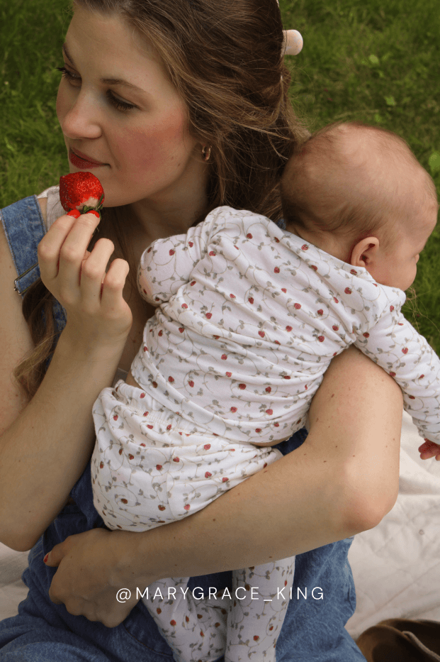 Woman holding a baby in a strawberry outfit while eating a strawberry outdoors. @marygraceking