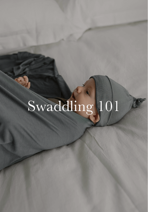 Infant swaddled in a gray stretchy fabric blanket with matching knotted hat lying on a white bed.