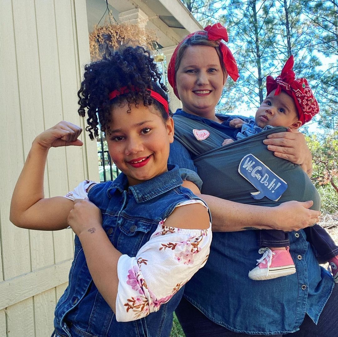 Girl in denim vest and floral top flexing arm, woman in denim shirt with red headscarf holding baby in gray baby carrier