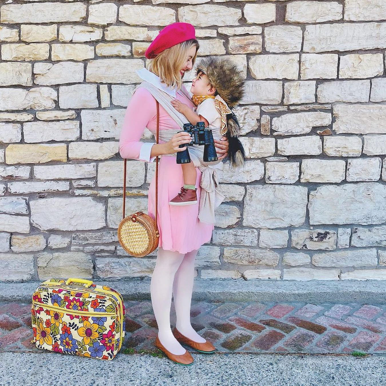 Woman in pink dress and red beret carrying baby with fur hat and binoculars in beige wrap carrier by stone wall with floral suitcase.