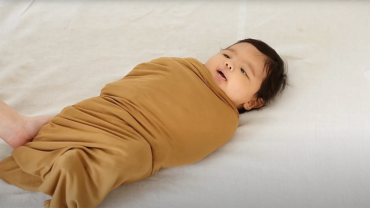 Baby swaddled in a mustard yellow stretch blanket lying on a light gray surface.
