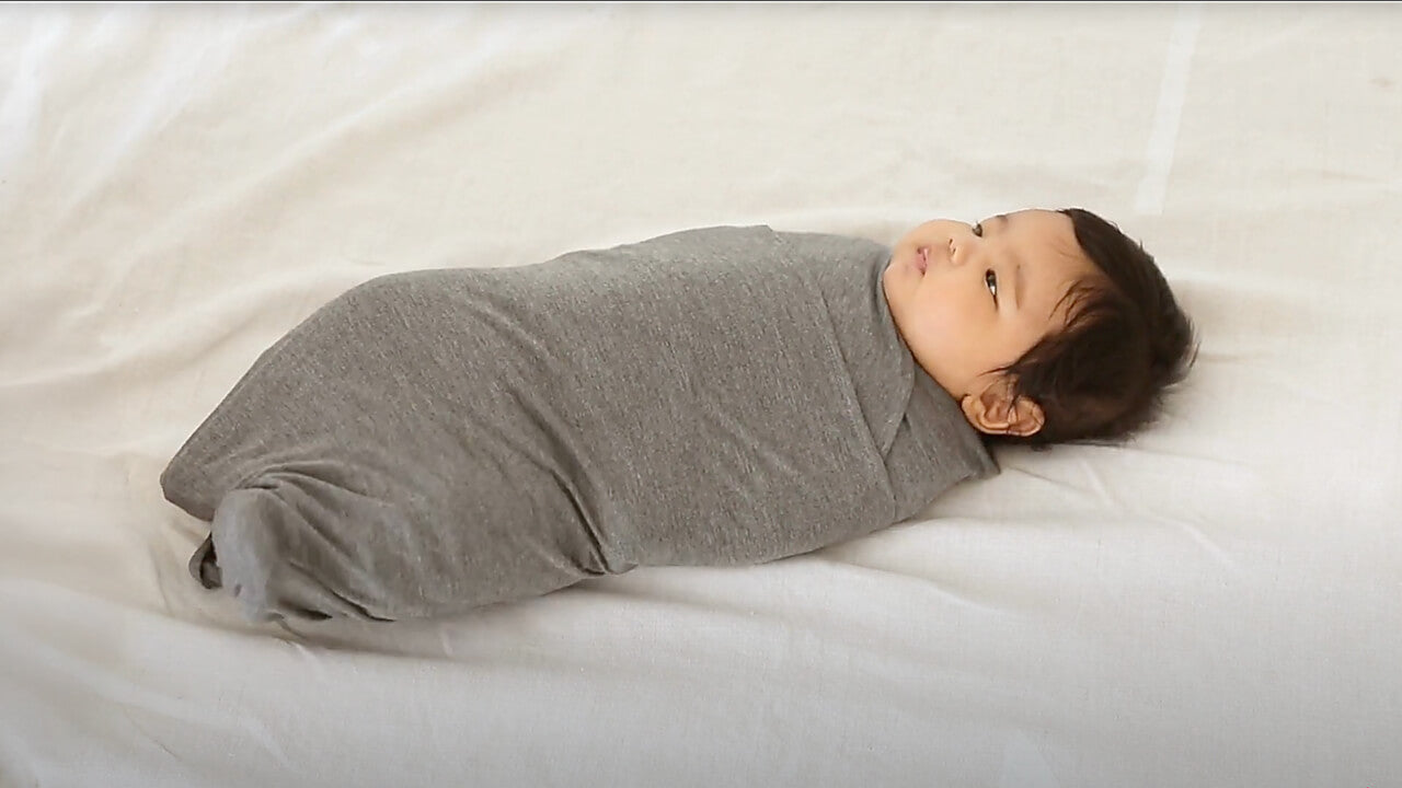 Baby wrapped snugly in a grey swaddle blanket lying on a white surface.