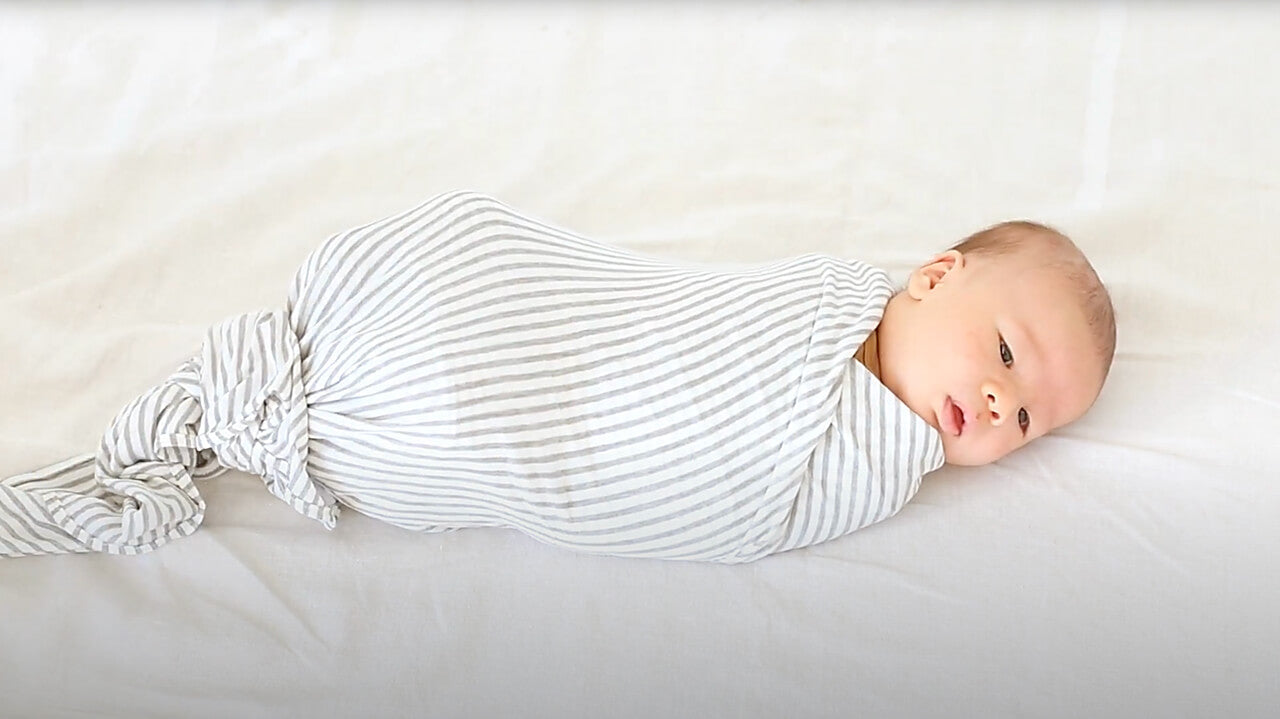 Baby swaddled in white and gray striped knot wrap blanket lying on white surface.