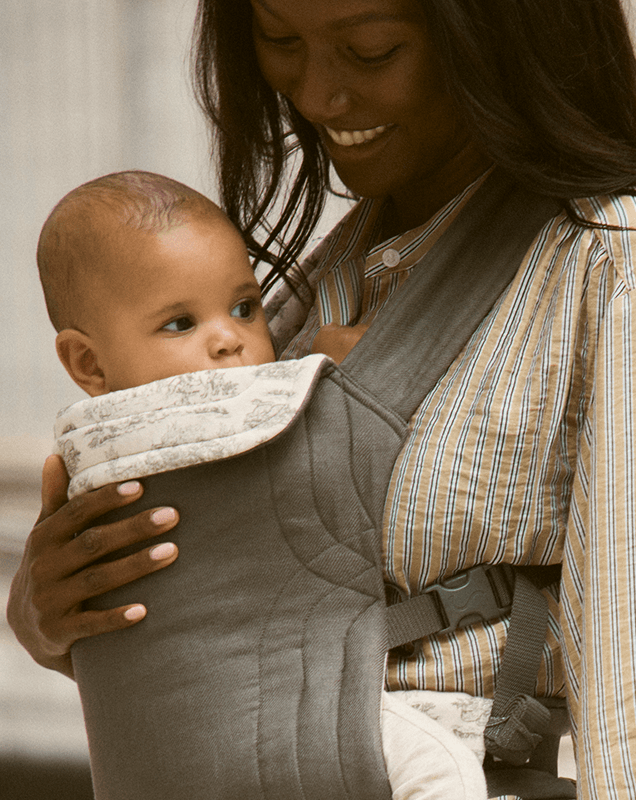 Dark gray fabric baby carrier with quilted detailing and an off-white patterned bib, worn by a woman in a striped shirt.