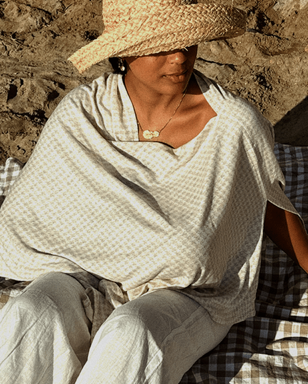 Woman wearing beige and white checkered poncho and straw woven hat sitting on matching checkered cushion outdoors.