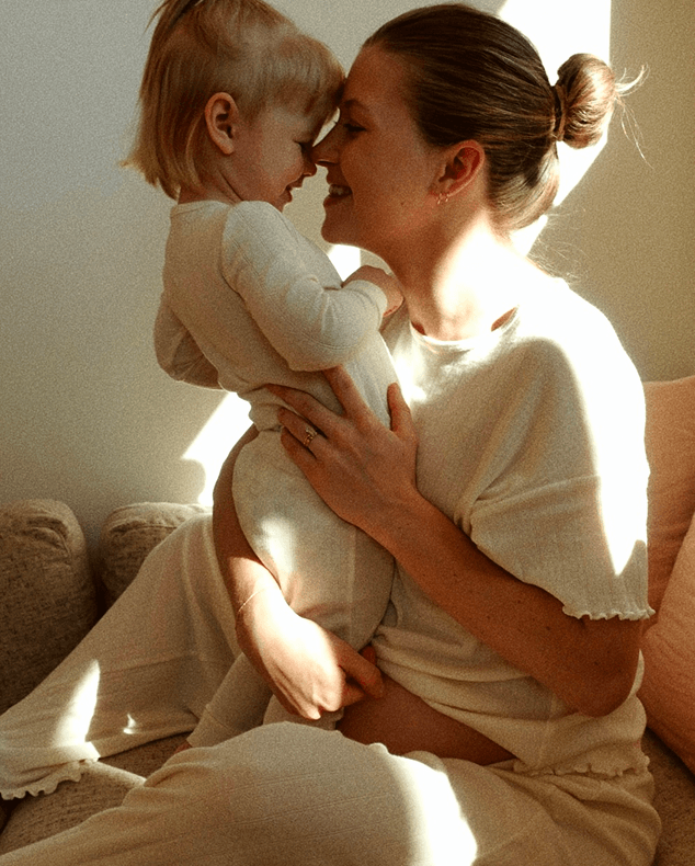 Woman in cream-colored loungewear holding a baby in white long-sleeve onesie, both smiling indoors on a couch.