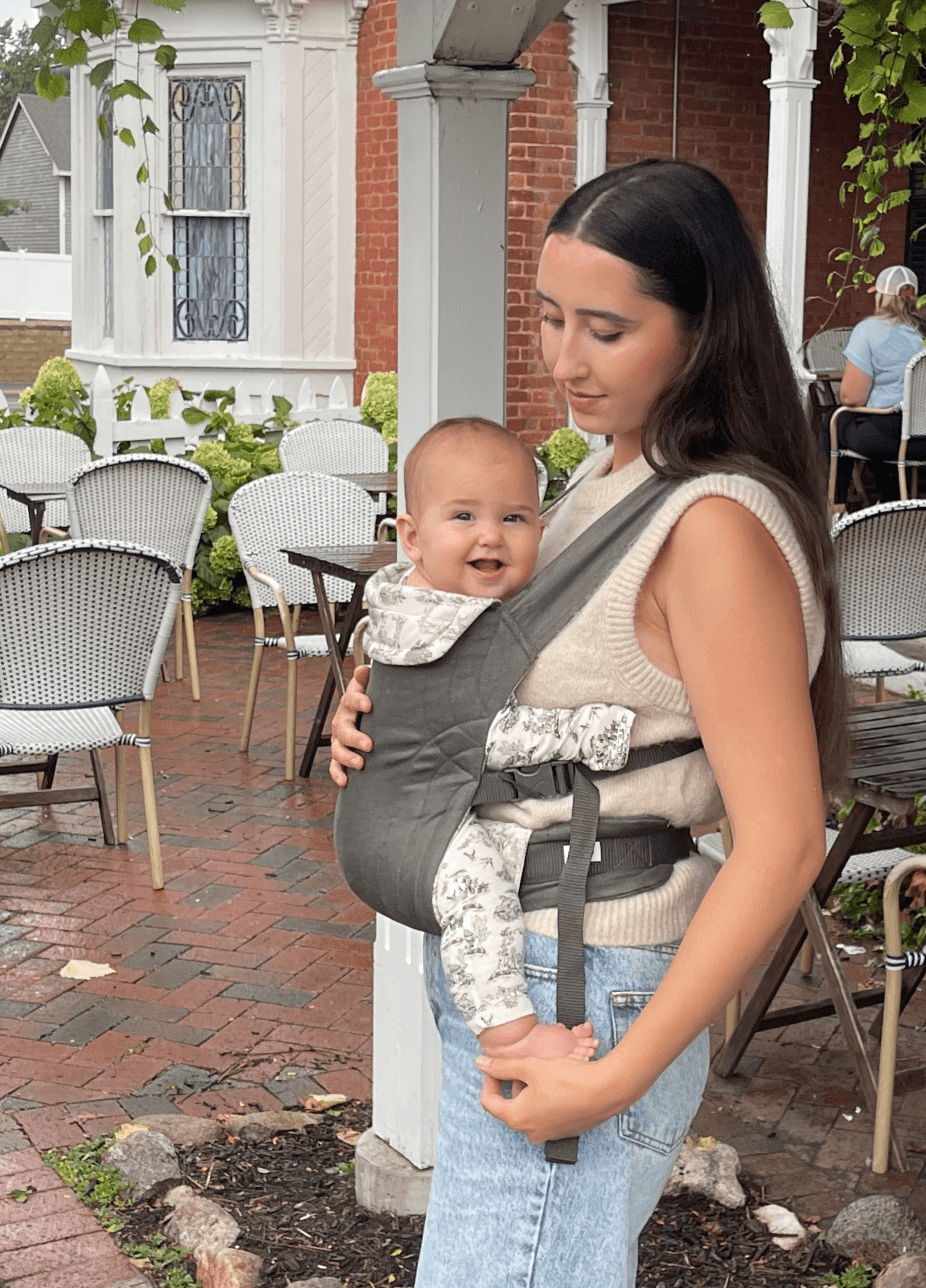 Woman wearing a gray baby carrier holding a smiling baby in white patterned clothing, standing outdoors by white chairs and brick flooring.