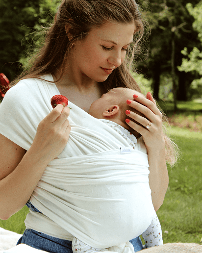 White stretchy baby wrap carrier holding infant close to mother's chest in outdoor park setting.