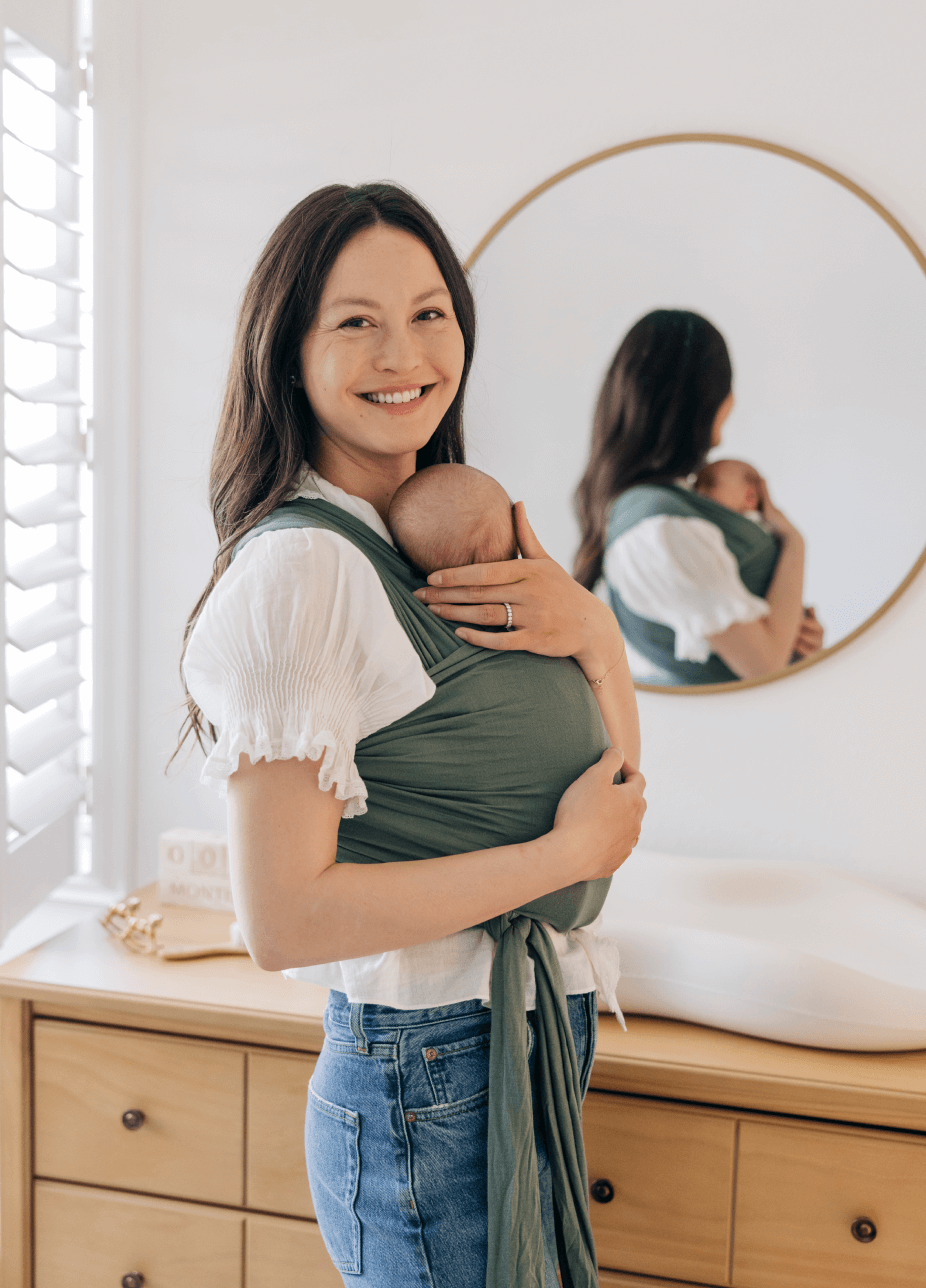 Woman smiling carrying baby in a green fabric baby wrap carrier indoors with mirror and wooden dresser background