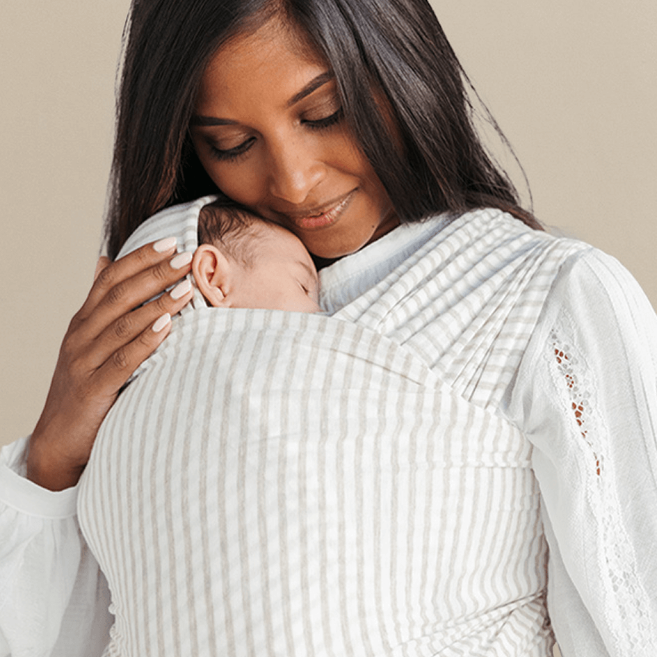 Beige and white striped baby wrap carrier holding sleeping baby, worn by person in white blouse with lace details.