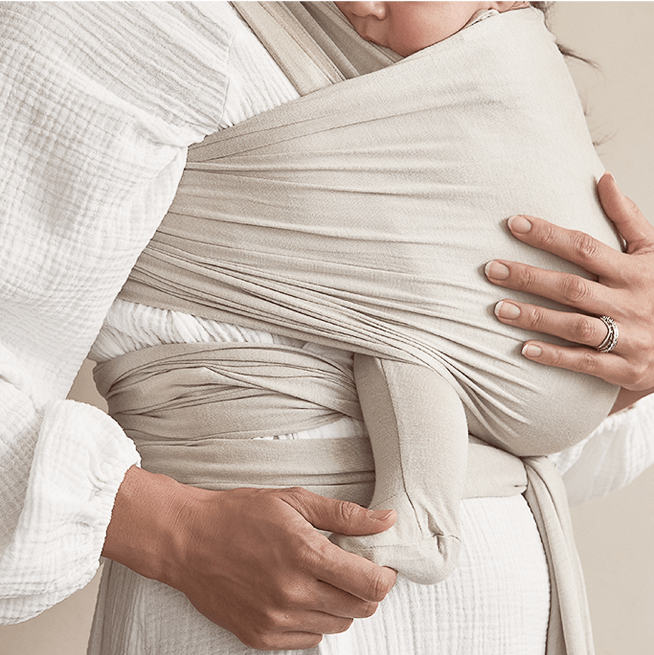 Beige fabric baby wrap carrier holding a baby dressed in beige leggings and white textured top.