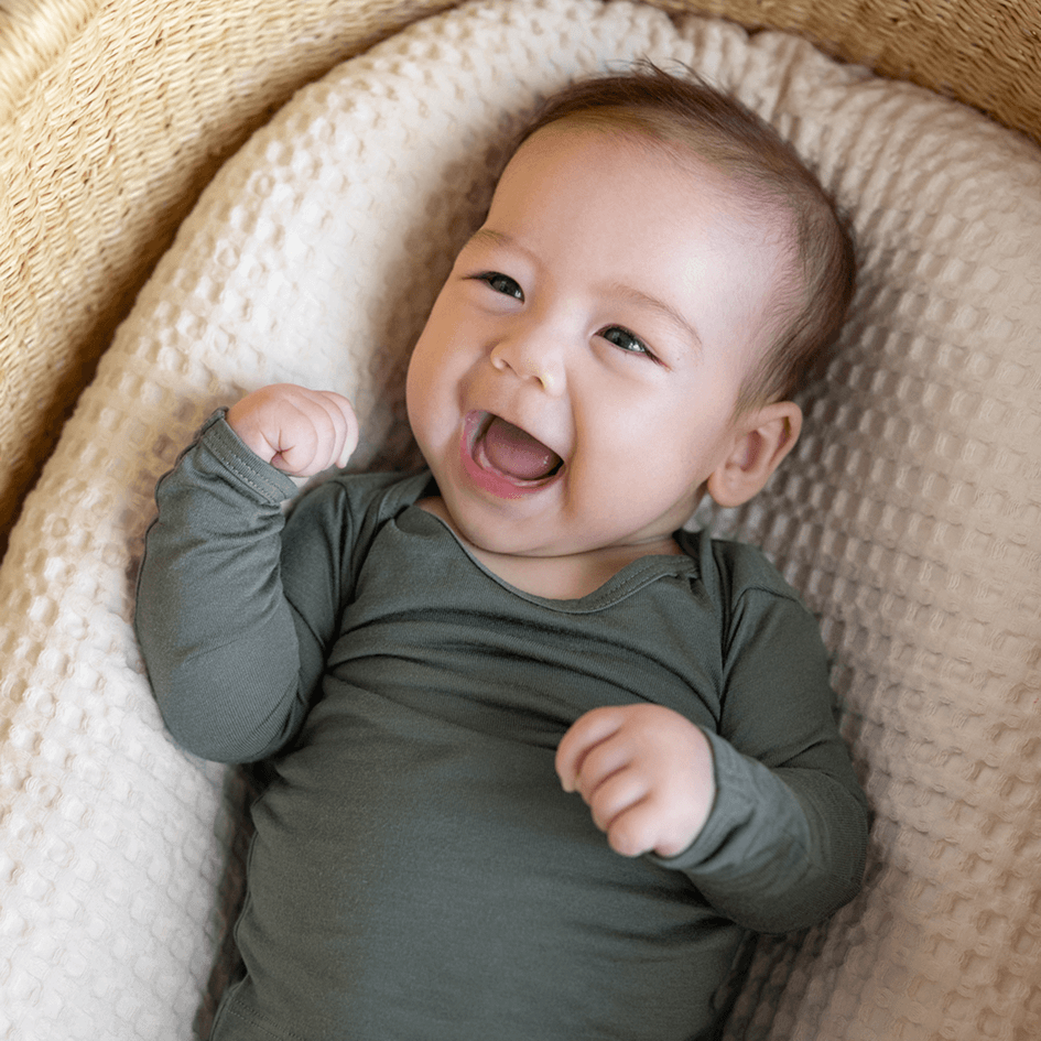Smiling baby lying on cream textured cushion wearing long-sleeve olive green onesie.