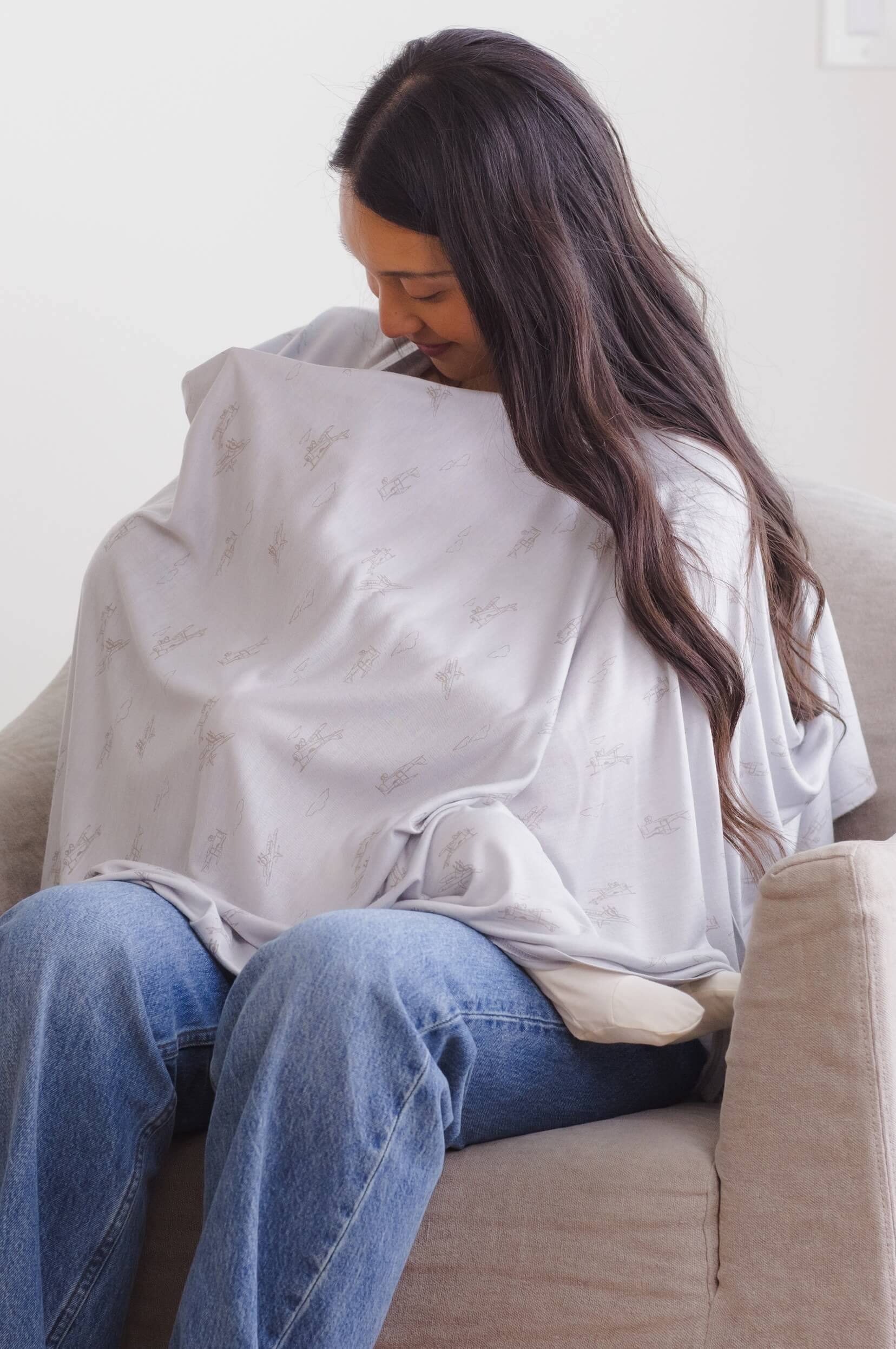 Woman using a light gray nursing cover with small airplane patterns while sitting on a beige sofa.