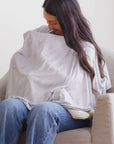 Woman using a light gray nursing cover with small airplane patterns while sitting on a beige sofa.