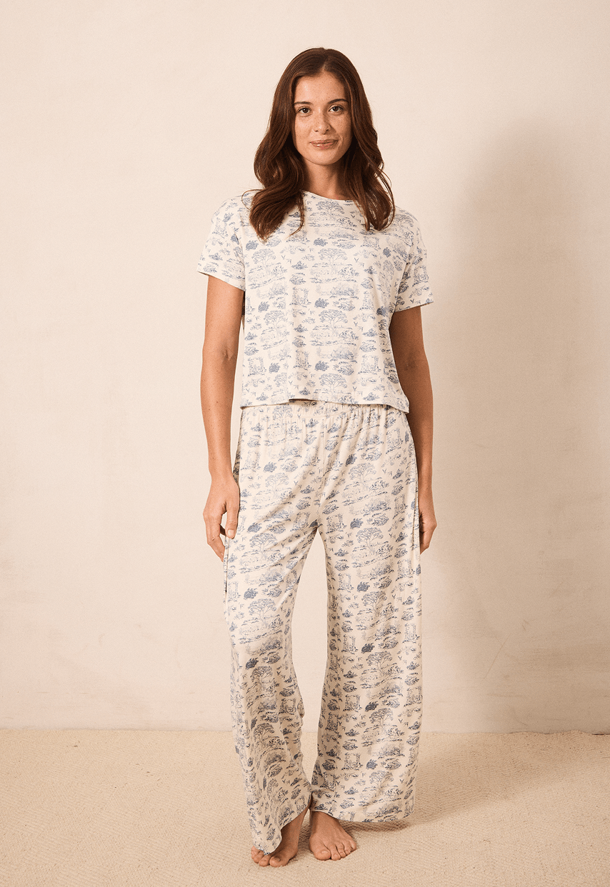 A woman modeling a matching two-piece pajama set with a short-sleeve top and wide-leg pants, featuring a light beige base with a blue toile pattern, set against a plain beige studio backdrop.