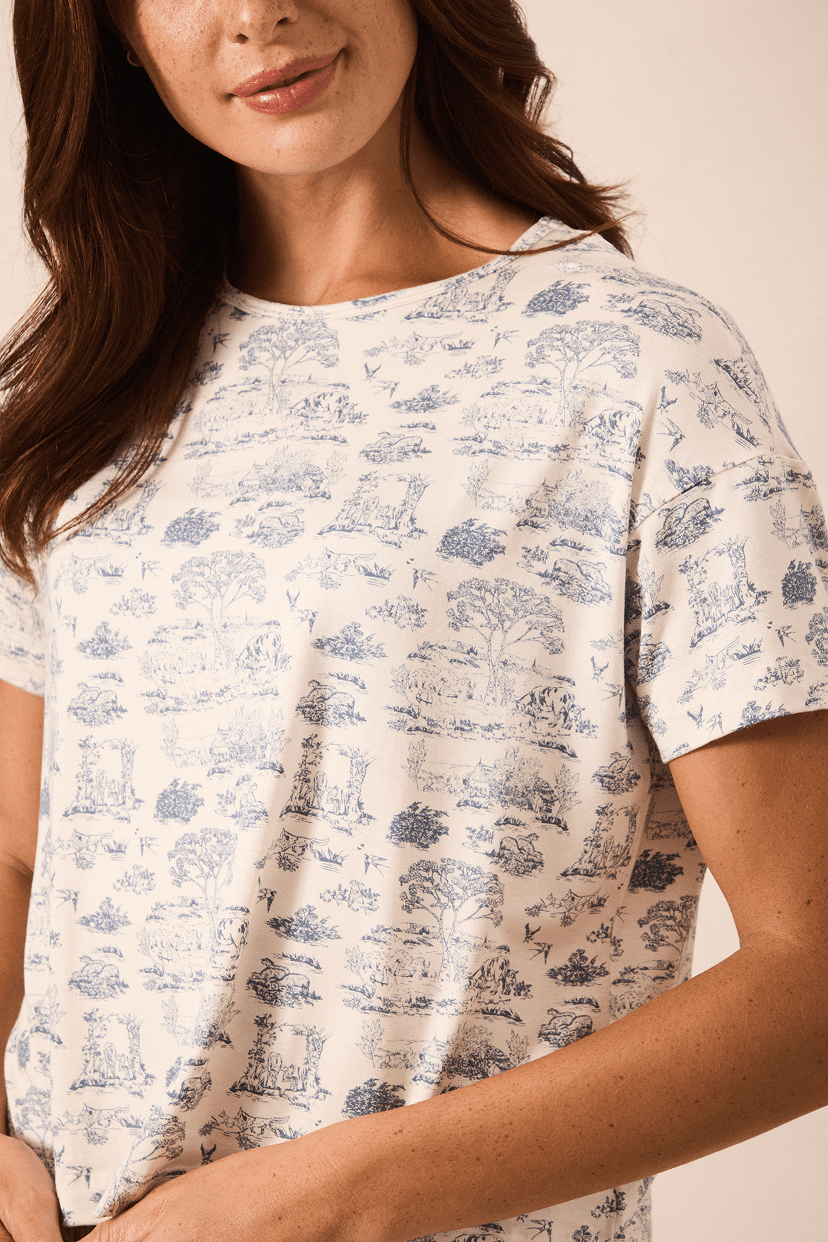 A close-up of a woman wearing a short-sleeve crewneck t-shirt with a blue toile de Jouy pattern on a cream-colored fabric, set against a warm beige background.