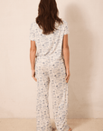 A woman viewed from behind wearing a matching short-sleeve top and wide-leg pants pajama set with a blue toile pattern on a cream background, standing against a plain beige wall.