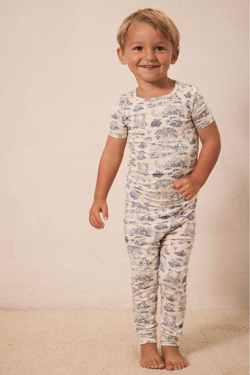 Toddler boy in cream-colored short sleeve top and pants with blue scenic print pajamas standing barefoot.