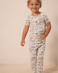 Toddler boy in cream-colored short sleeve top and pants with blue scenic print pajamas standing barefoot.