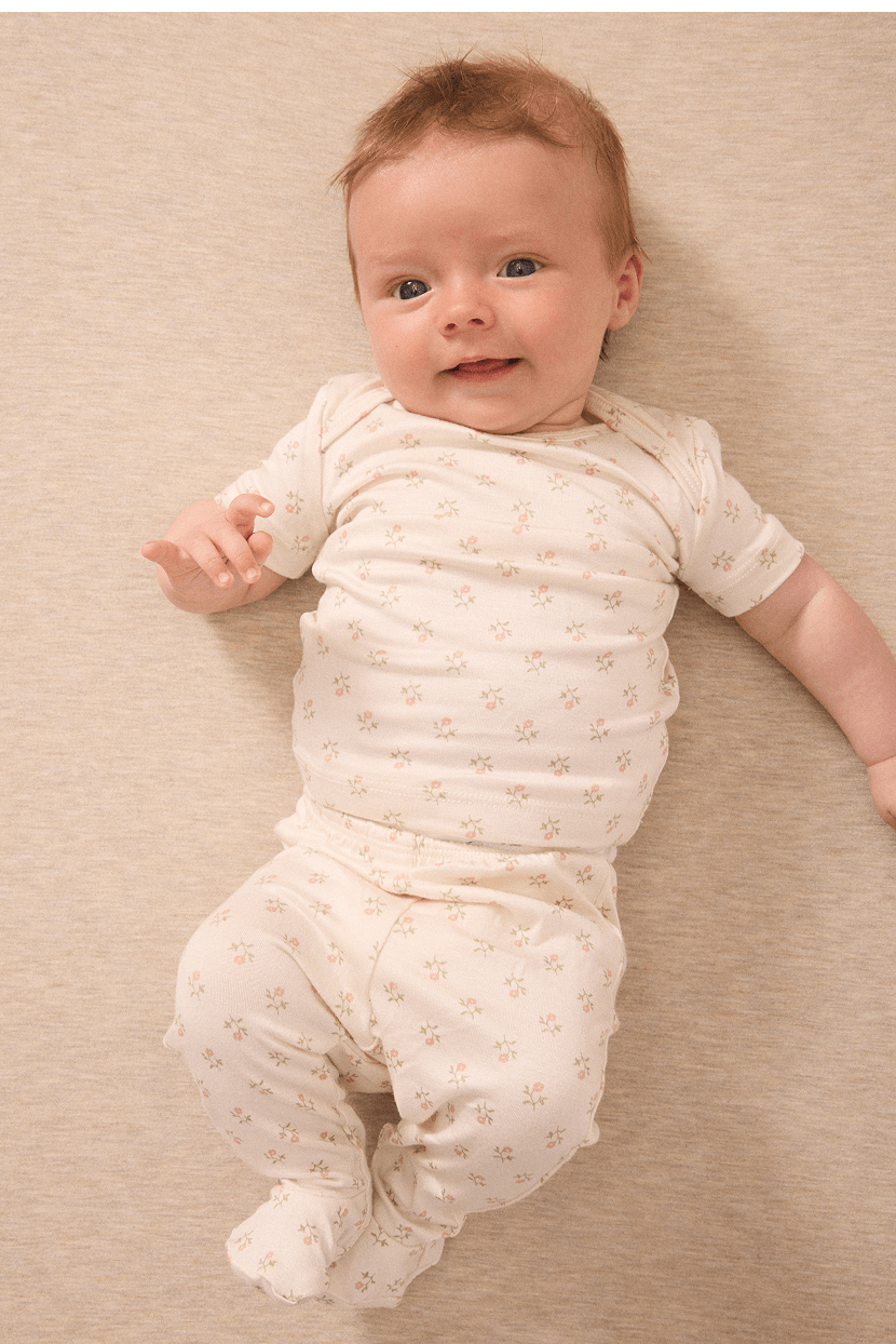 Baby wearing cream-colored two-piece outfit with small pink floral print, lying on beige fabric background.