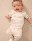 Baby wearing cream-colored two-piece outfit with small pink floral print, lying on beige fabric background.