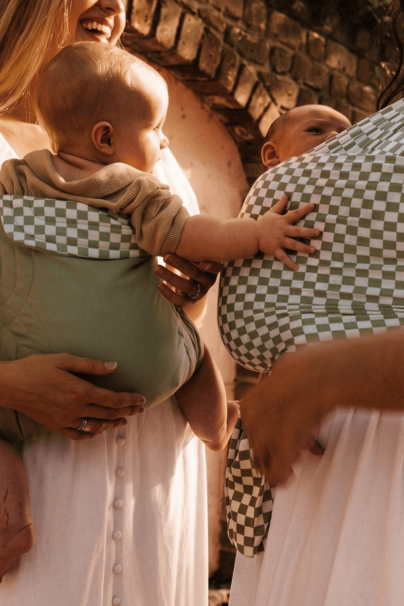Two babies carried in olive green and green checkered baby carriers worn by adults in white dresses.