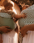 Two babies carried in olive green and green checkered baby carriers worn by adults in white dresses.
