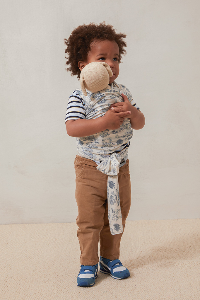 Toddler wearing brown pants, striped shirt, blue sneakers, and a blue and white patterned fabric wrap holding a beige plush toy.