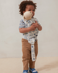 Toddler wearing brown pants, striped shirt, blue sneakers, and a blue and white patterned fabric wrap holding a beige plush toy.
