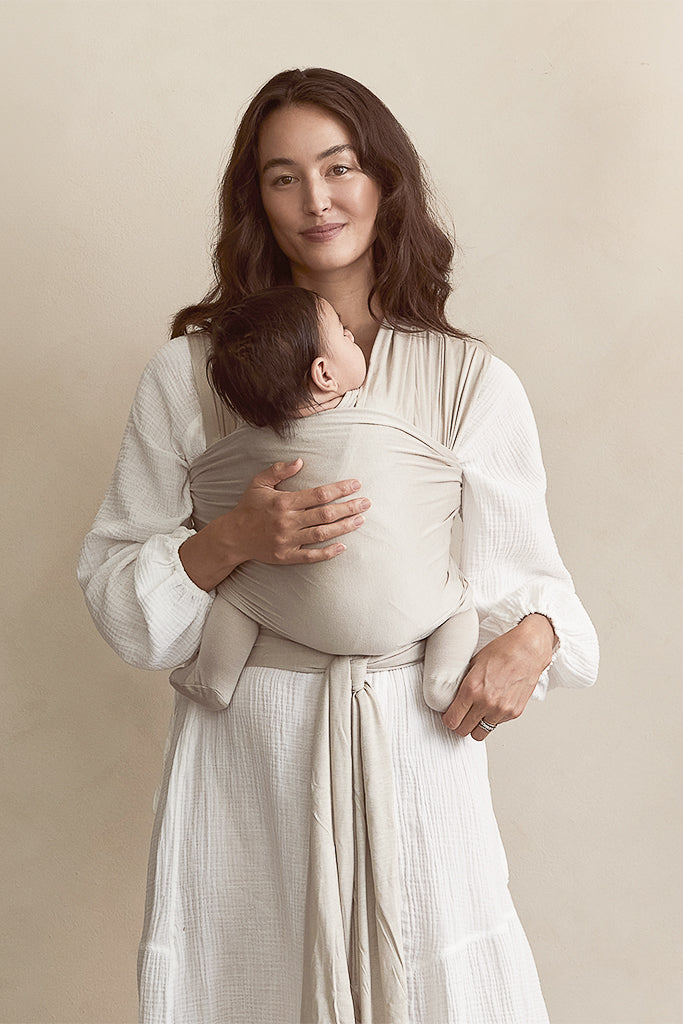 Woman wearing a white textured dress carrying a baby in a beige baby wrap carrier tied at the waist.