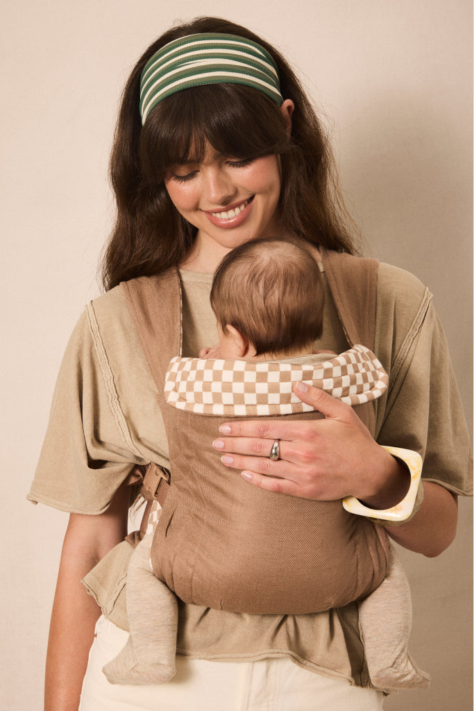A woman wearing a beige baby carrier wrap holds a baby in a front-facing baby carrier. The carrier has a solid beige body with a cream and beige checkered lining on the shoulder straps.