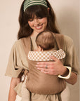 A woman wearing a beige baby carrier wrap holds a baby in a front-facing baby carrier. The carrier has a solid beige body with a cream and beige checkered lining on the shoulder straps.